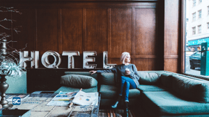 A Woman Sitting On A Couch With Coffee In A Hotel Lobby With A Shoppers Confidential Logo